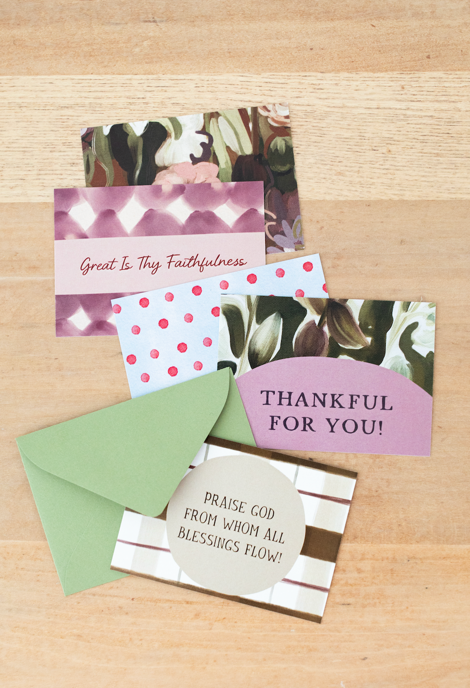 A set of handmade thank-you cards and envelopes arranged on a light wood background. The cards feature various patterns including pink and white diamonds, pink and white polka dots, green and white leaf prints, and brown and white stripes, with inspirational religious quotes printed on them.