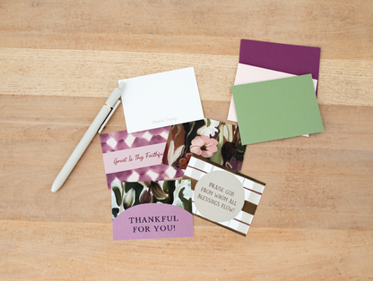 A flat-lay of various paper craft or stationery items on a light wood background, including patterned cards, a white envelope, a white pen, and colored paper sheets.
