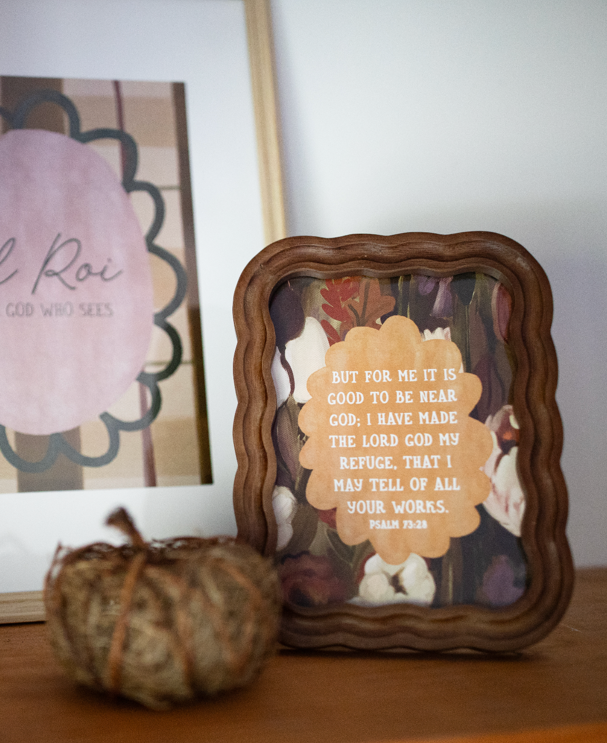 A framed decorative art print with a scalloped brown wooden frame sits on a wooden surface. The print features a beige flower-shaped label with the text of Psalm 34:18 over a background of white doves and autumn leaves.