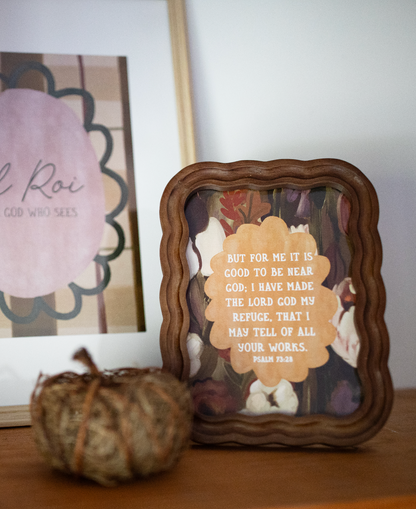 A framed decorative art print with a scalloped brown wooden frame sits on a wooden surface. The print features a beige flower-shaped label with the text of Psalm 34:18 over a background of white doves and autumn leaves.