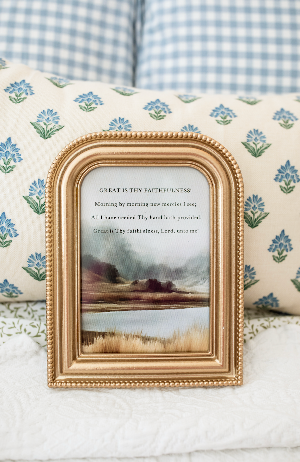 A small gold-framed art print with a religious poem and a landscape painting, styled on a white quilted blanket in front of floral pillows.