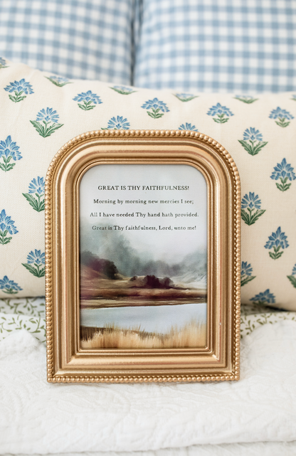 A small gold-framed art print with a religious poem and a landscape painting, styled on a white quilted blanket in front of floral pillows.