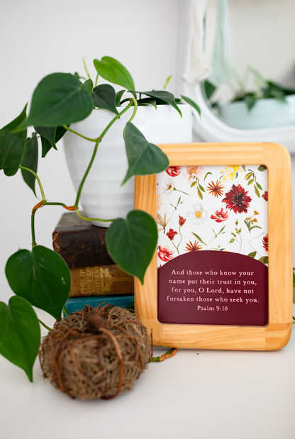 A square wooden-framed Bible verse plaque with a floral print background and a dark red text panel reading Psalm 9:10, styled on a white surface with a green plant and decorative books behind it.