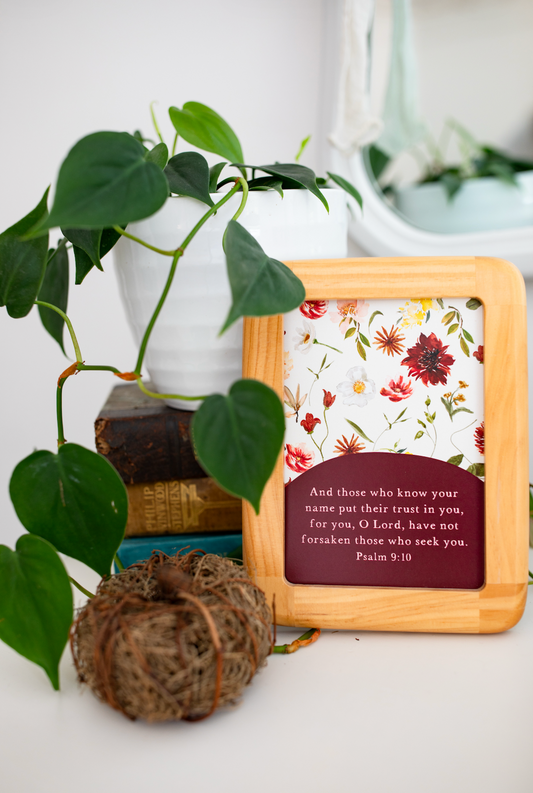 A square wooden-framed Bible verse plaque with a floral print background and a dark red text panel reading Psalm 9:10, styled on a white surface with a green plant and decorative books behind it.