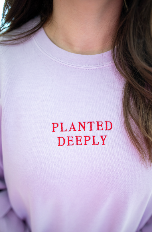 Close-up of a person wearing a light lavender crewneck sweatshirt with 'PLANTED DEEPLY' embroidered in red across the chest.
