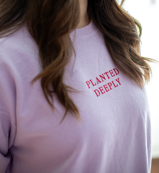 Close-up of a person wearing a light lavender crewneck sweatshirt with 'PLANTED DEEPLY' embroidered in red across the chest.