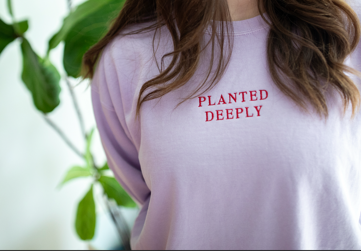 A close-up of a person wearing a light lavender crewneck sweatshirt with 'PLANTED DEEPLY' printed in red text on the chest, with green leaves blurred in the background.