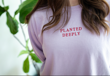 A close-up of a person wearing a light lavender crewneck sweatshirt with 'PLANTED DEEPLY' printed in red text on the chest, with green leaves blurred in the background.